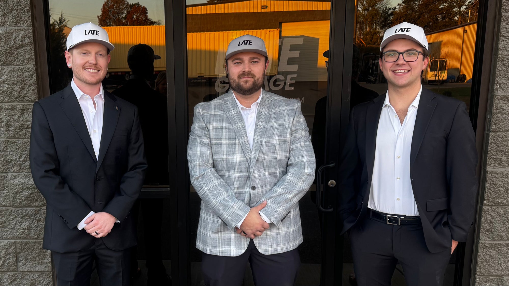 Three men standing in front of a glass door wearing matching white baseball caps with 'LATE' printed on the front, along with business attire.